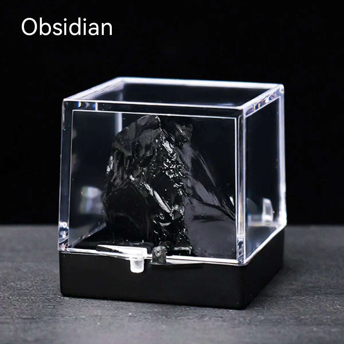 A clear glass box containing a dark obsidian rough stone specimen on a black surface.