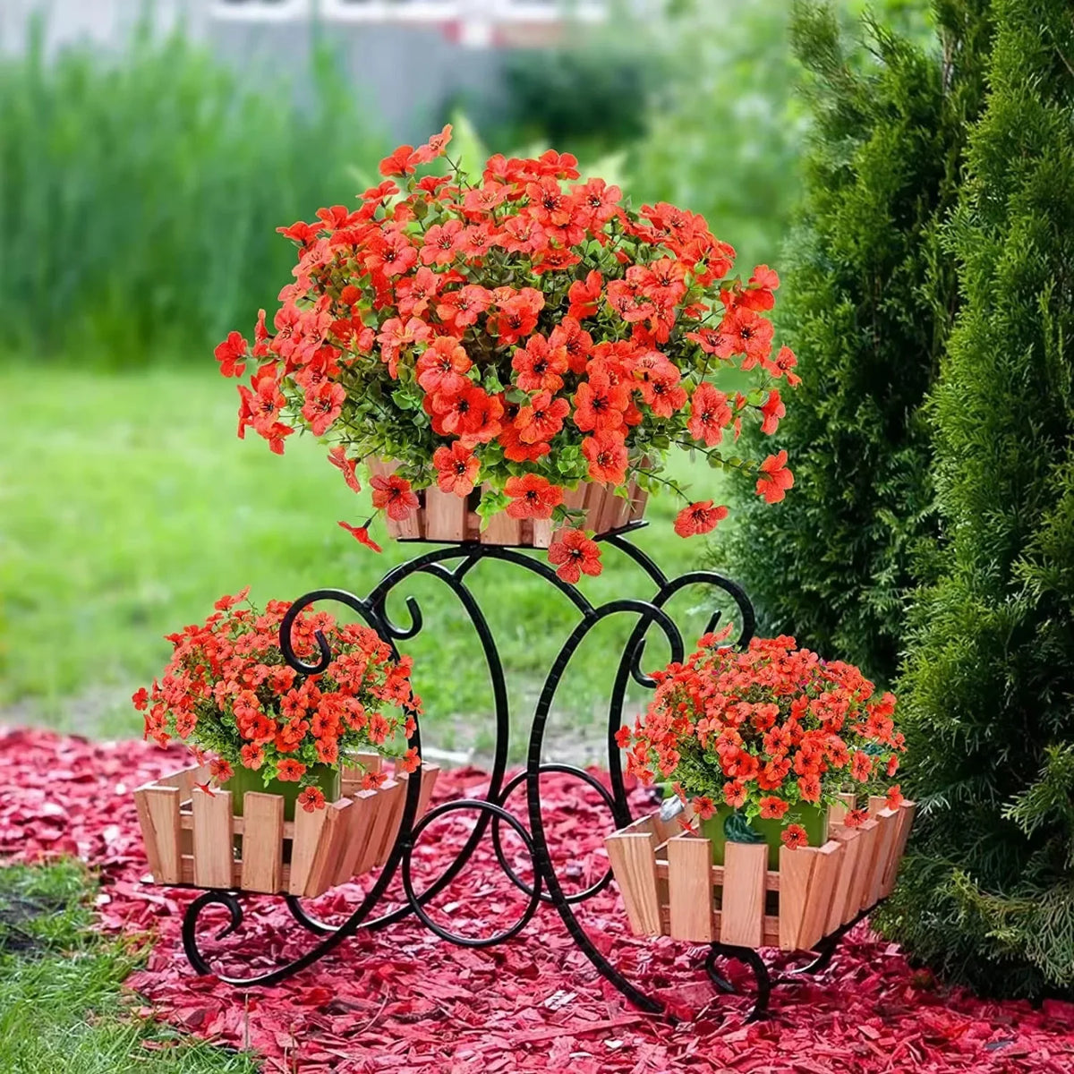 A display of bright orange and red artificial flowers in wooden planters mounted on a black metal stand, set against green fo