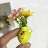 A hand holds a small yellow ceramic vase with red spots and artificial pink and yellow flowers.