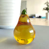 A clear glass figurine shaped like a pear with a green top, resting on a light surface.
