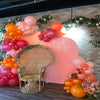 A pink and orange balloon garland decoration featuring balloons, flowers, and a wicker chair against a brick wall.