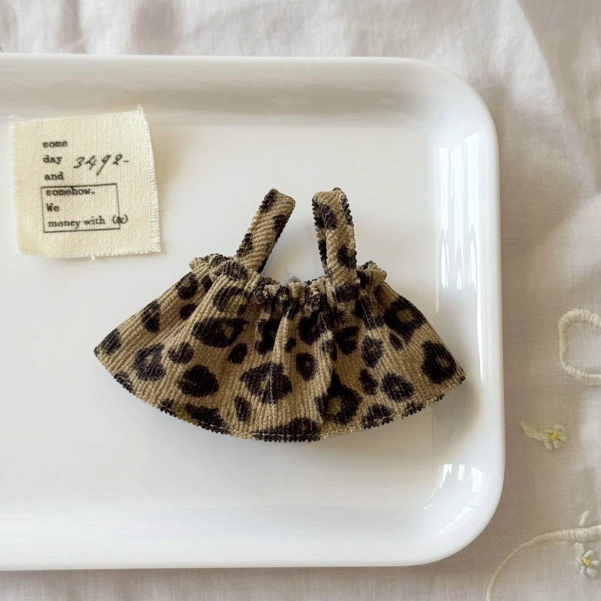 A leopard print shoulder strap skirt for dolls is displayed on a white plate next to a piece of paper.