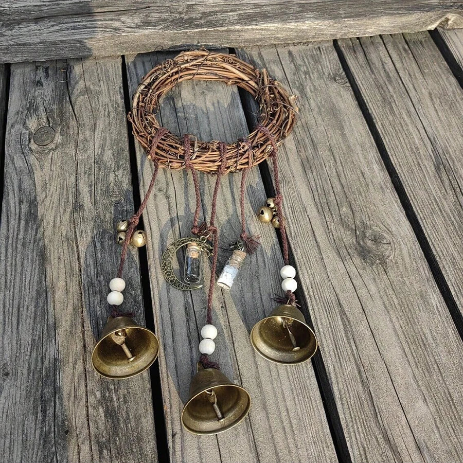 A rustic wreath of twigs with hanging brass bells and leather cords on a weathered wooden surface.