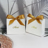 Two white paperboard thank you gift bags tied with gold ribbon resting on a white surface.
