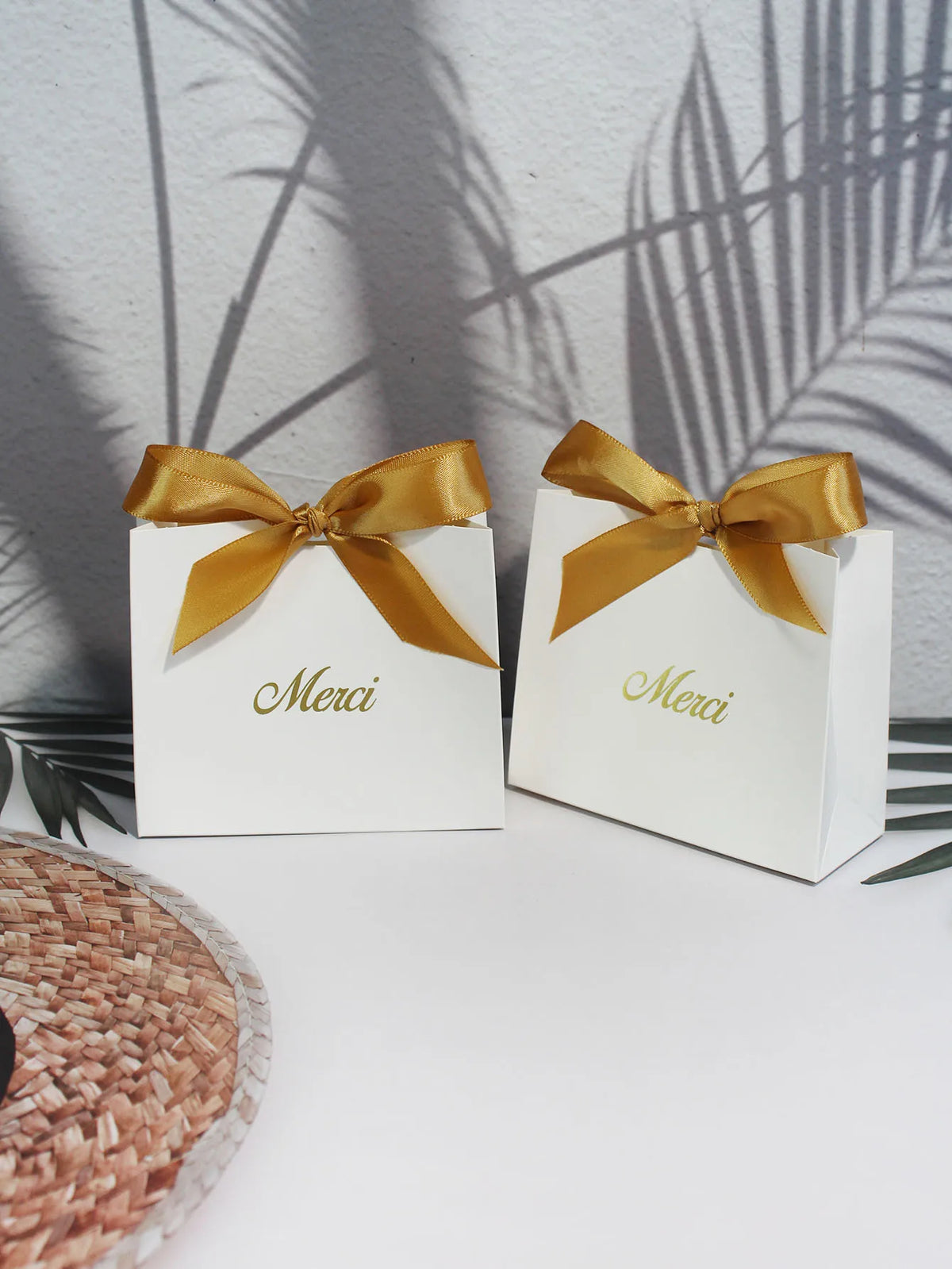 Two white paperboard thank you gift bags tied with gold ribbon resting on a white surface.