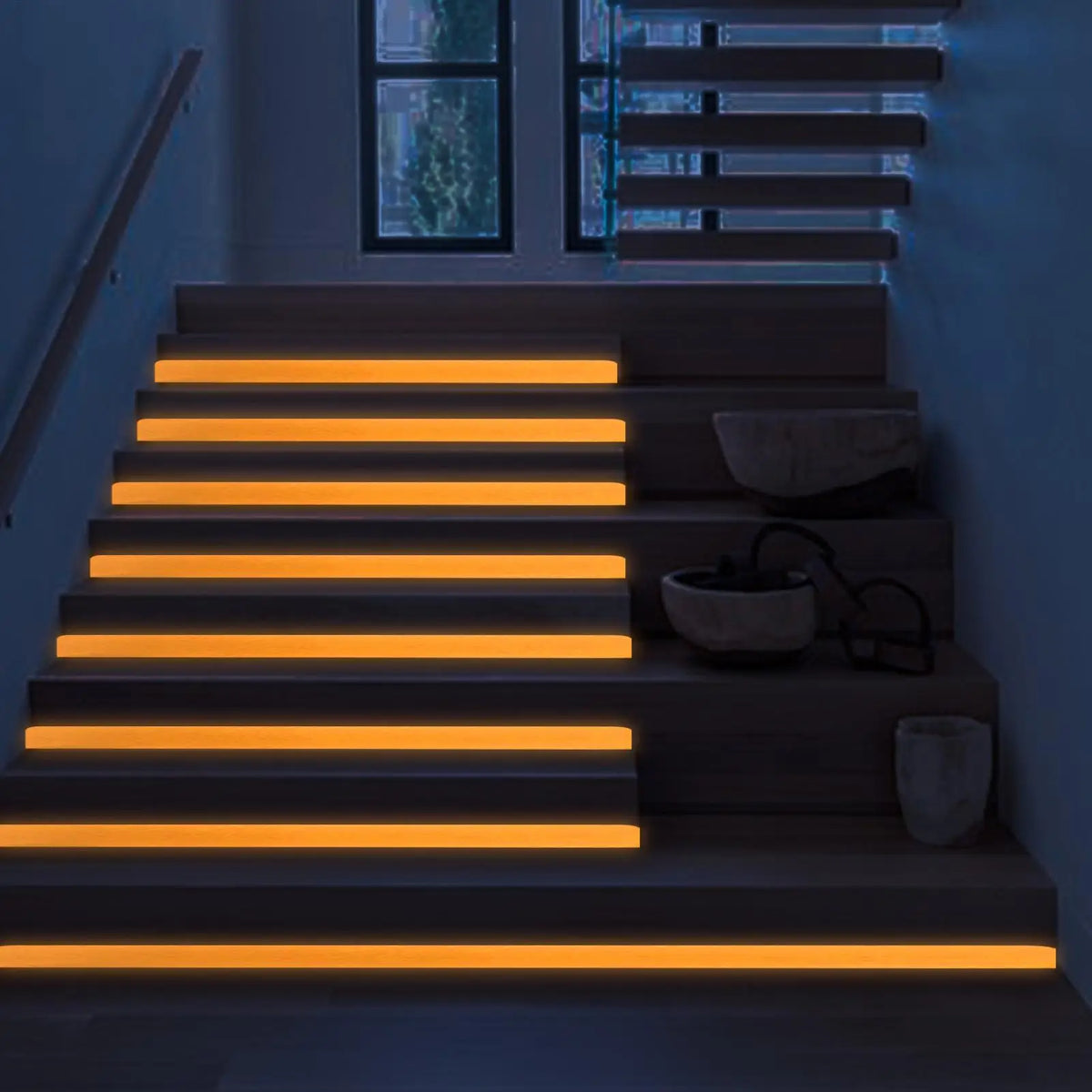 A modern staircase illuminated by warm orange LED strips, highlighting the steps and a window view.