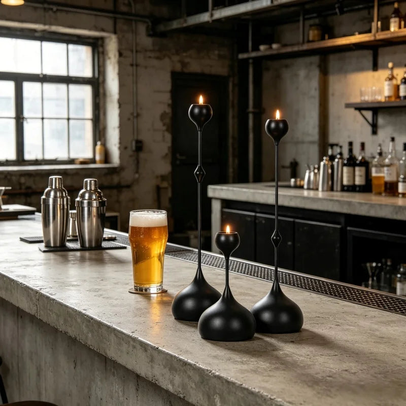 A set of three black candle holders with lit candles resting on a rustic bar counter in an industrial setting.