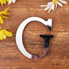 A silver acrylic letter G wall sticker placed on a wooden surface with yellow and white flowers.