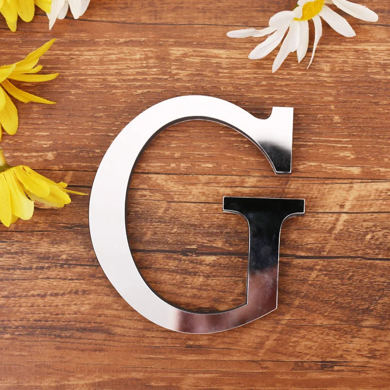 A silver acrylic letter G wall sticker placed on a wooden surface with yellow and white flowers.