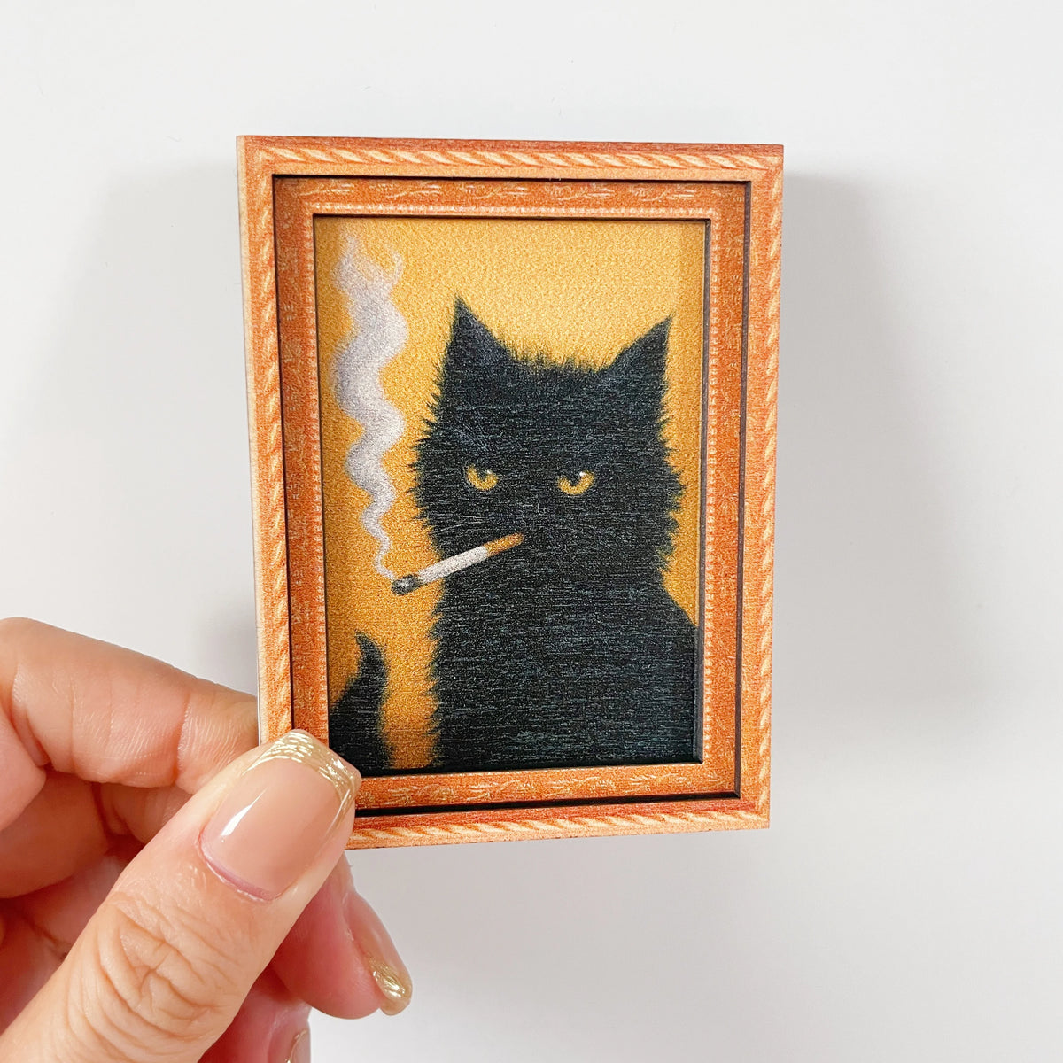 Una mano sostiene una foto enmarcada de un gato negro con humo saliendo de su nariz, diseñada como imanes para nevera.