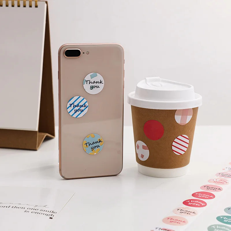 Ein beige Smartphone zeigt runde Dankessticker auf der Rückseite neben einer braunen Papptasse für Kaffee.