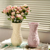 A white ceramic vase with floral relief patterns sits on a white cloth next to a pink textured vase.