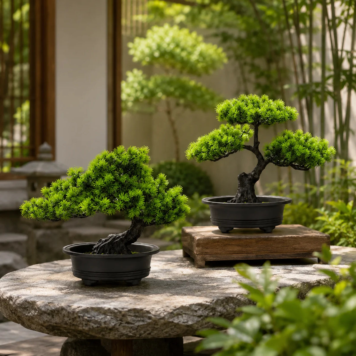An artificial bonsai pine tree in a black pot sits on a stone surface surrounded by lush greenery.