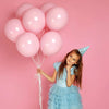 A young girl in a light blue dress and a cone hat holds strings of pink balloons against a pink background.