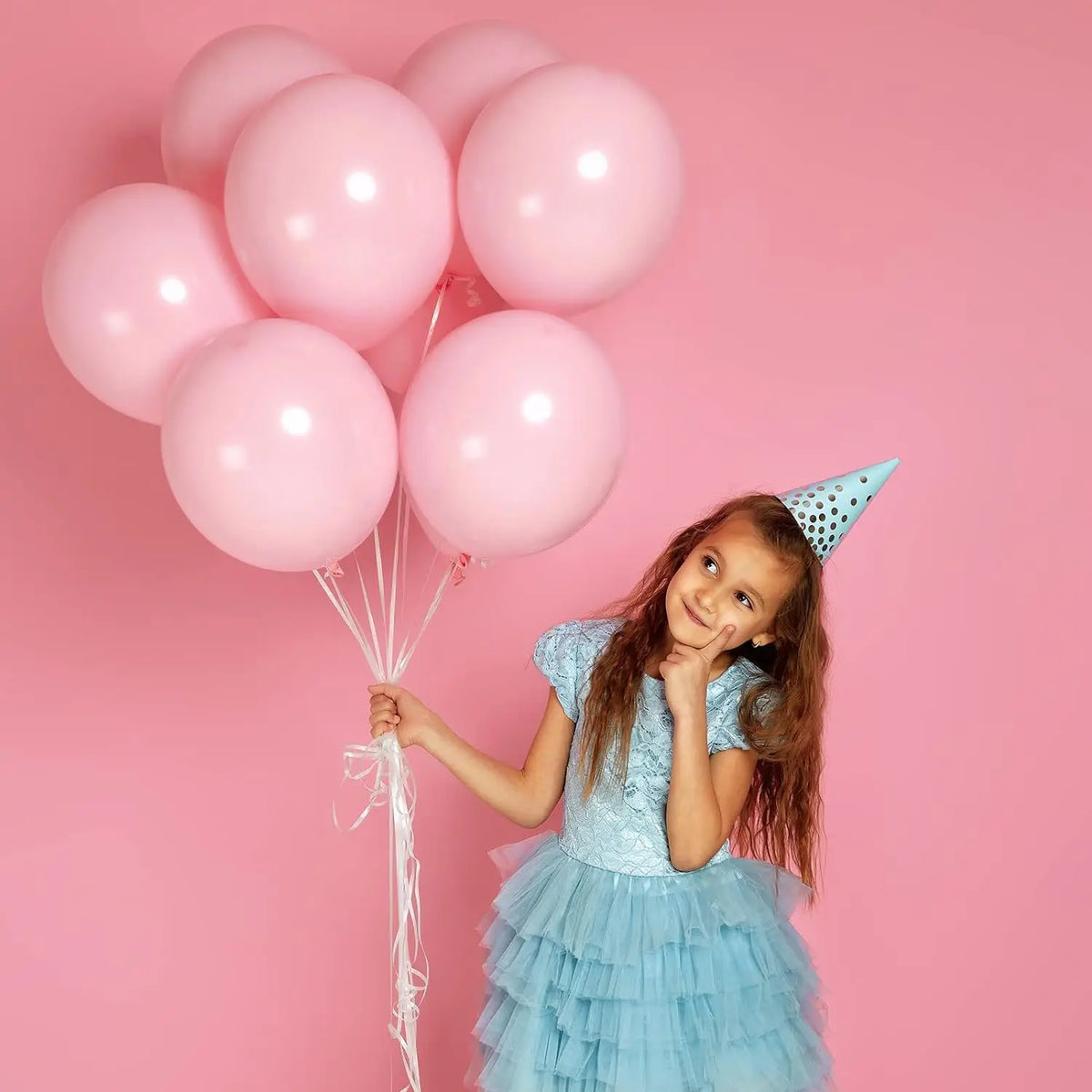 A young girl in a light blue dress and a cone hat holds strings of pink balloons against a pink background.