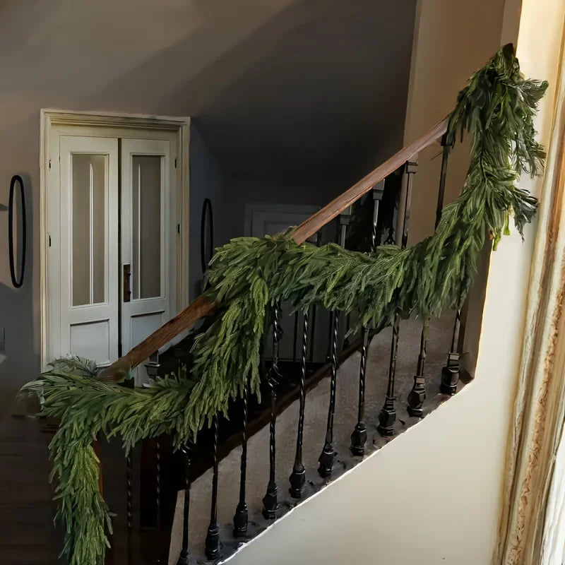 An artificial Norfolk pine garland with warm white lights draped along a wooden railing.