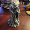 A dark gray sculpture of a Grim Reaper couple embracing, displayed on a wooden surface.