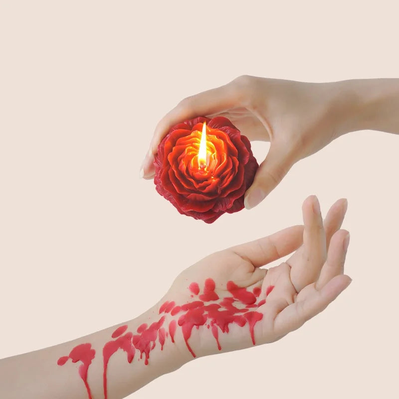 A hand holds a glowing red rose-shaped candle, with red paint splatters on the palm of another hand.