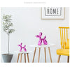 Pink resin balloon dog sculpture on a white table against a white wall with a yellow chair and a potted plant.