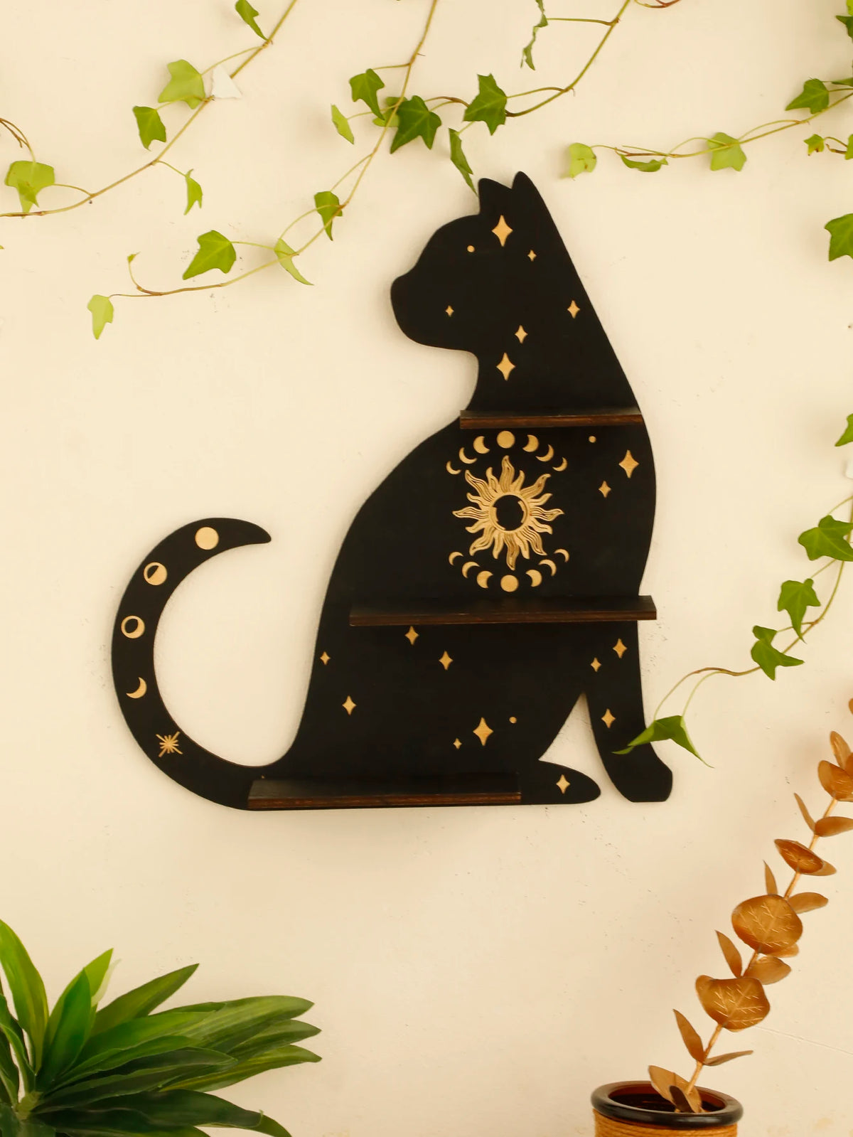 A black silhouette of a cat with gold accents and stars, mounted on a wall shelf surrounded by green vines.