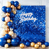 A collection of gold and blue balloons arranged around a bright blue sequin wall backdrop with Happy Birthday text.