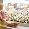 A young girl with blonde hair is sitting by a window looking at a field of white and yellow daisy flowers.