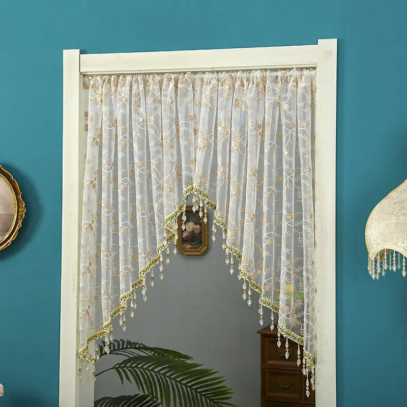A sheer lace door curtain with gold tassels hangs in a white frame against a teal wall.