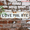 A distressed white metal sign with the text LOVE YOU BYE mounted on a brick wall.
