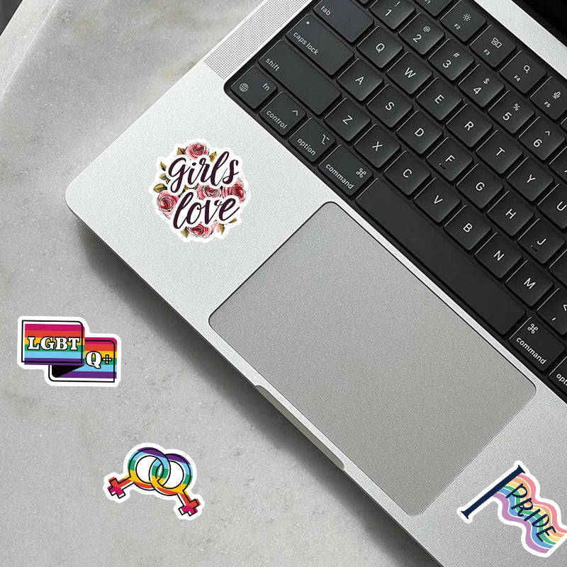 A laptop keyboard and touchpad display various LGBTQ pride and love themed stickers on a gray background.