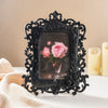 A black ornate resin photo frame with a square glass insert rests on a white surface next to a cream-colored fabric.