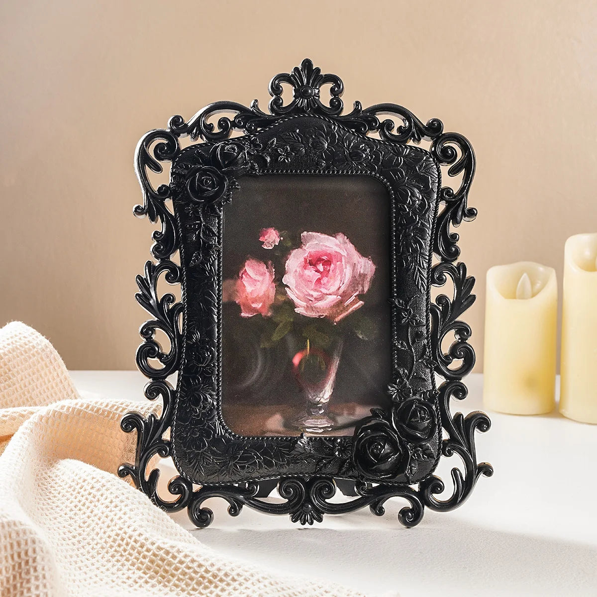 A black ornate resin photo frame with a square glass insert rests on a white surface next to a cream-colored fabric.