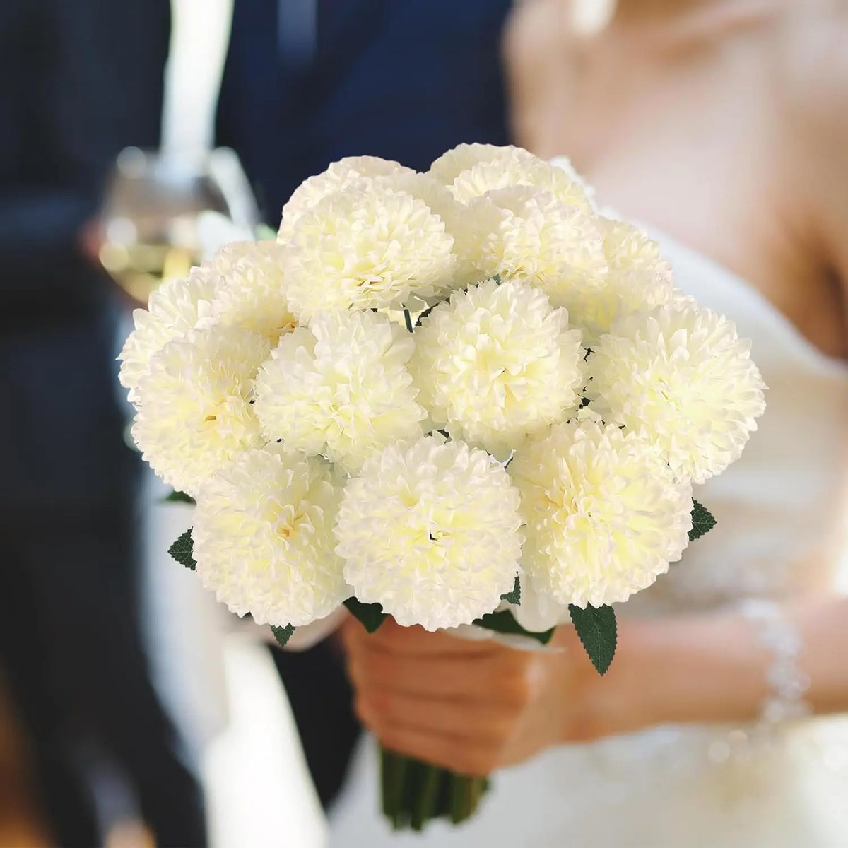 A bouquet of white artificial chrysanthemum flower balls held by a person in a wedding setting.