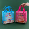 A hand holds a pink reusable happy birthday gift bag with a cake on top against a green background.