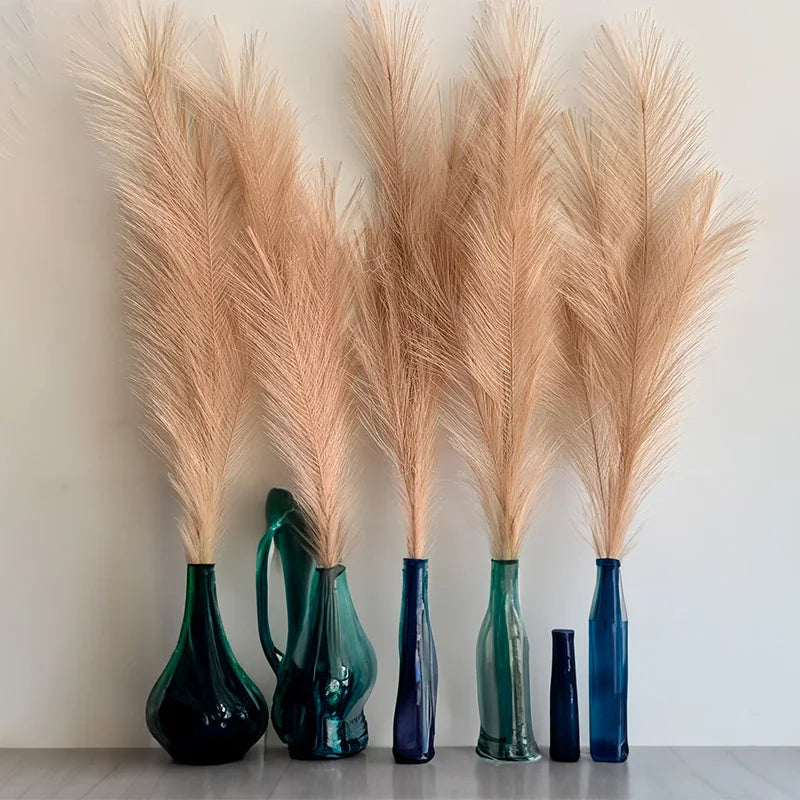 A collection of tall, dried pampas grass stems in various shades of beige and pink displayed against a white wall.