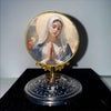 A round religious ornament featuring a depiction of the Virgin Mary with folded hands against a light background.