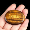 A polished, oval amber crystal with the word Courage engraved on its surface is held in a person's hand.