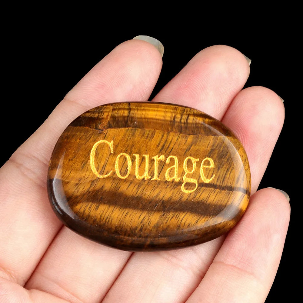 A polished, oval amber crystal with the word Courage engraved on its surface is held in a person's hand.