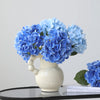 A bouquet of artificial blue hydrangea flowers in a white ceramic vase on a dark surface.