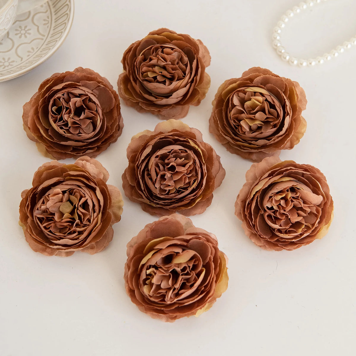A collection of dried silk peony artificial flowers in shades of dusty rose and brown arranged on a white surface.