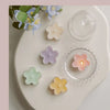 A white ceramic plate displays a handmade soy wax flower tea light candle in various pastel colors and a glass lid.