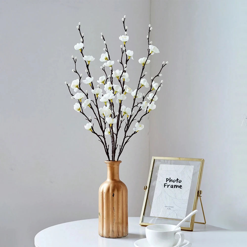 A vase holds artificial plum blossom decorations with bare branches on a white surface.