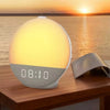 A modern sunrise alarm clock with a glowing white light and digital display sits on a wooden surface against a sunset backgro