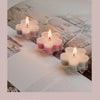 A close-up of a handmade soy wax flower tea light candle with a lit flame set in a glass holder on a white surface.