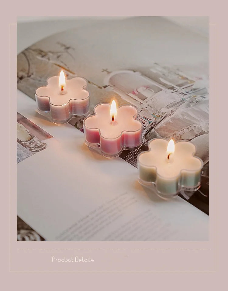 A close-up of a handmade soy wax flower tea light candle with a lit flame set in a glass holder on a white surface.