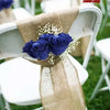 A close-up of a white chair decorated with a burlap ribbon tied around the back, featuring a dark blue artificial rose.