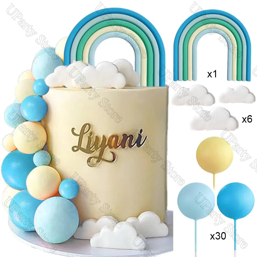 A white cake decorated with blue and yellow ball and cloud toppers, featuring a rainbow arch topper.