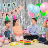 A group of children celebrating a birthday in front of a silver metallic foil fringe curtain backdrop with colorful balloons 