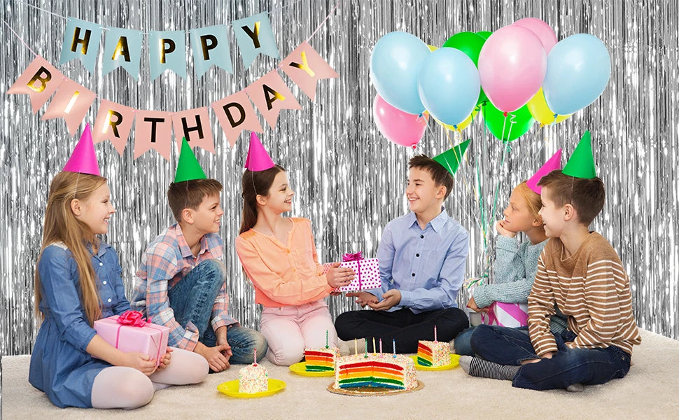 A group of children celebrating a birthday in front of a silver metallic foil fringe curtain backdrop with colorful balloons 