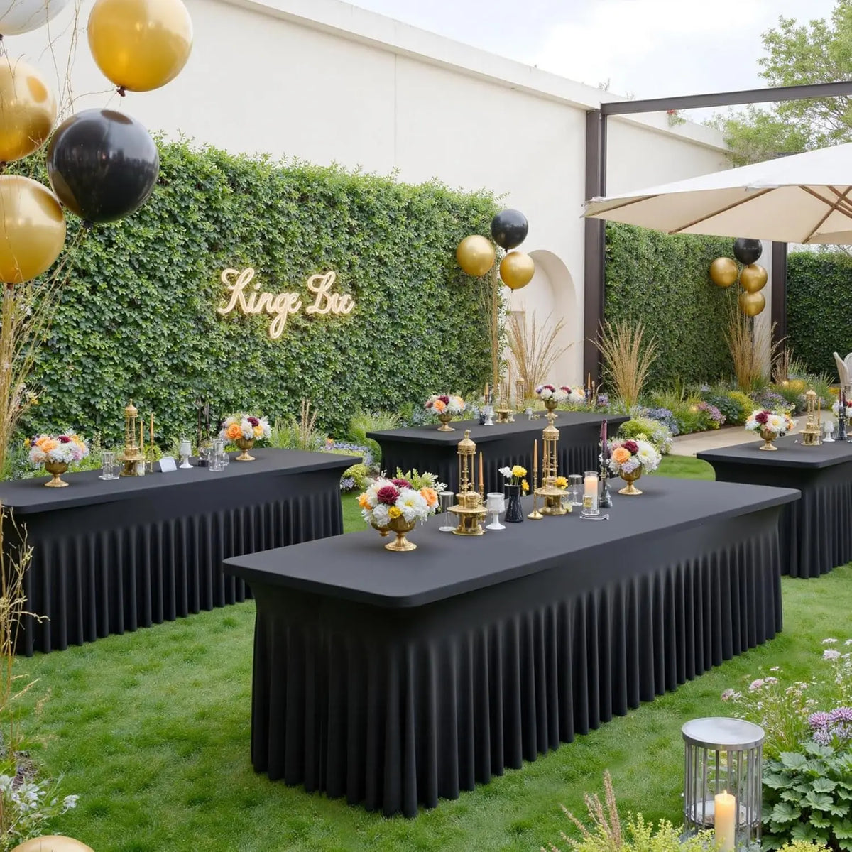 A long black pleated spandex tablecloth cover set up outdoors for an event with greenery and gold balloons.