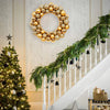 A golden and champagne silk Christmas ball wreath ornament hangs above a white staircase decorated with green foliage.